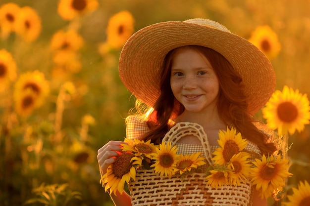 Happy life in summer portrait of a beautiful redhaired girl in a hat on a field of sunflowers with flowers in her hands in the rays of the setting sun