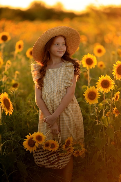 Happy life in summer portrait of a beautiful redhaired girl in a hat on a field of sunflowers with flowers in her hands in the rays of the setting sun