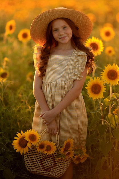 Happy life in summer portrait of a beautiful redhaired girl in a hat on a field of sunflowers with flowers in her hands in the rays of the setting sun