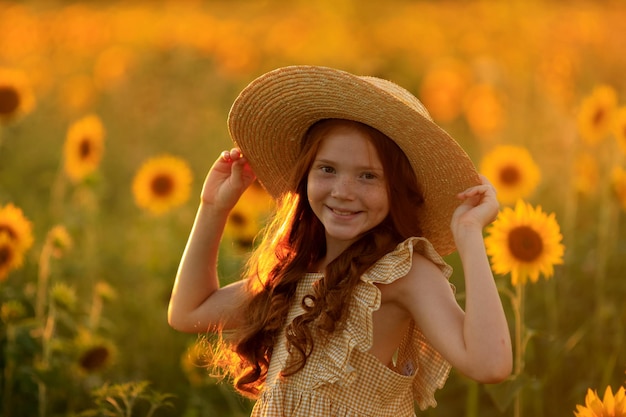 Happy life in summer portrait of a beautiful redhaired girl in a hat on a field of sunflowers with flowers in her hands in the rays of the setting sun