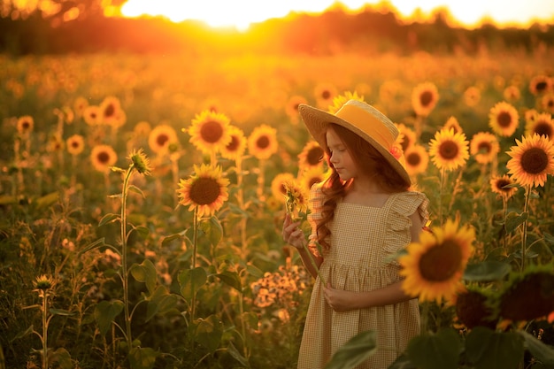 Happy life in summer portrait of a beautiful redhaired girl in a hat on a field of sunflowers with flowers in her hands in the rays of the setting sun