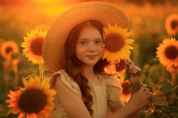 Happy life in summer portrait of a beautiful redhaired girl in a hat on a field of sunflowers with flowers in her hands in the rays of the setting sun