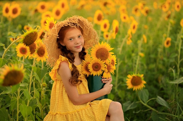 Happy life in summer portrait of a beautiful redhaired girl in a hat on a field of sunflowers with flowers in her hands in the rays of the setting sun