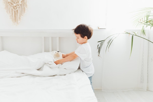 Happy little Asian boy cleans his room and folds a blanket in the bedroom at home The kid makes the bed adjusts the blanket on the big bed