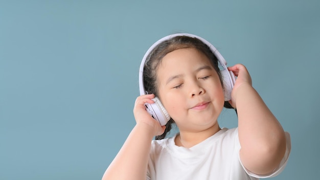 Happy little asian child girl  listening music in headphones over blue background. music, technology and people concept.