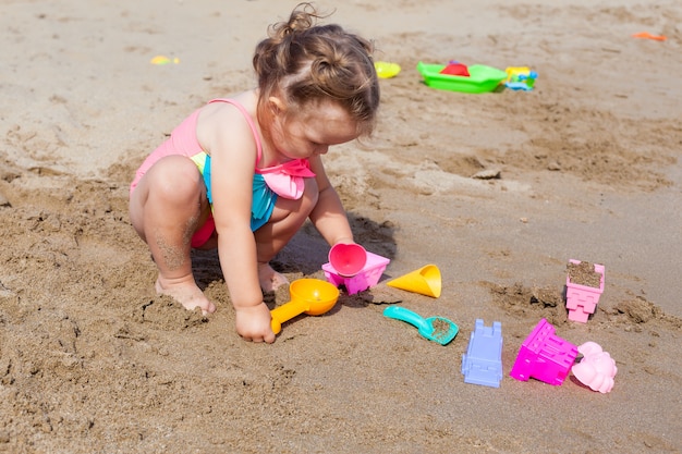 Happy little baby girl in swimsuit playing in the sand on the beach on a sunny warm day.