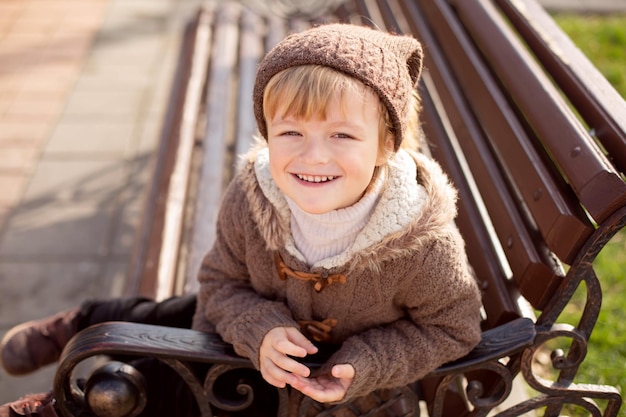 a happy little blond boy in knitted brown clothes is sitting on a park bench