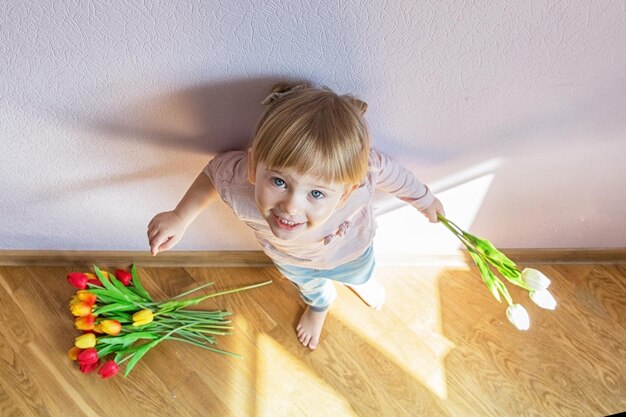 Happy little blond girl stands leaning against the wall in a sunny room at home