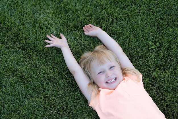 Happy little blond girl in a Tshirt lying on a green grass Smiling child is resting Concept of a carefree childhood Longawaited summer summer holidays