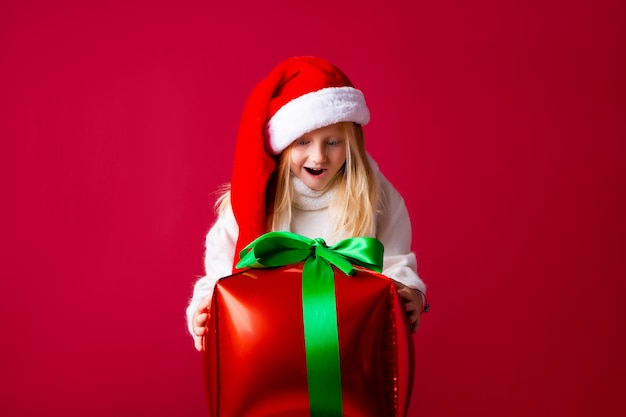 Happy little blonde girl in a santa hat holding a New Year's gift on a red wall