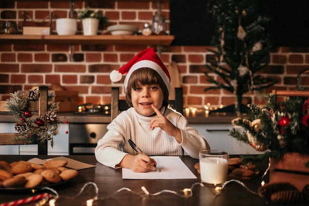 A happy little boy in a white knitted sweater and a red hat is sitting at a table and writing a New Year's letter