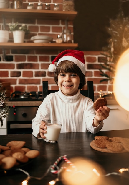 A happy little boy in a white sweater and a red Christmas hat is sitting at the kitchen table with oatmeal cookies with milk