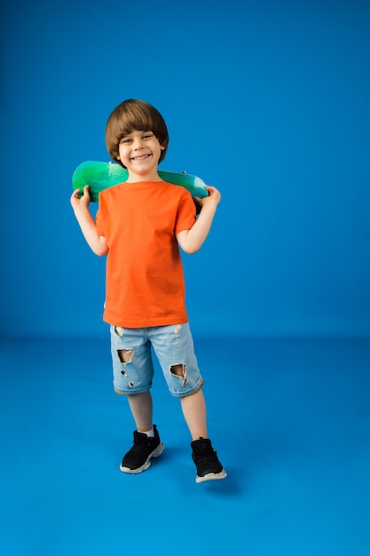 Happy little boy with brown hair holds a skateboard on a blue surface with space for text