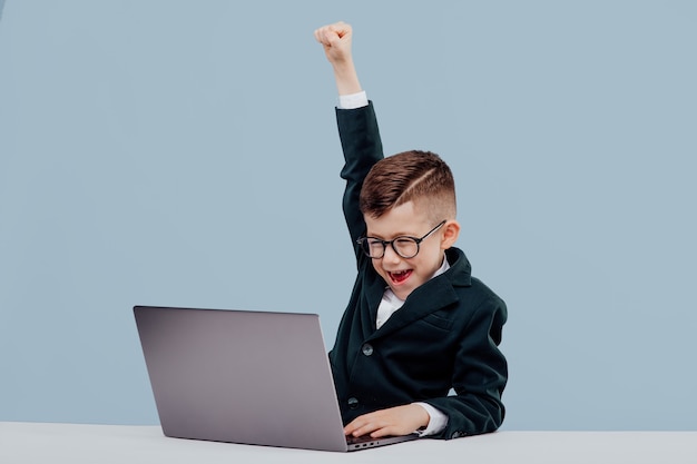 Happy little business boy enjoys victory hands up and glasses smiling and approving work in office a...