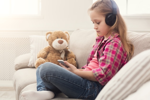 Happy little casual girl in headphones listening to music on smartphone. Pretty kid at home watching cartoon on mobile phone online, sitting on sofa with her teddy bear, copy space