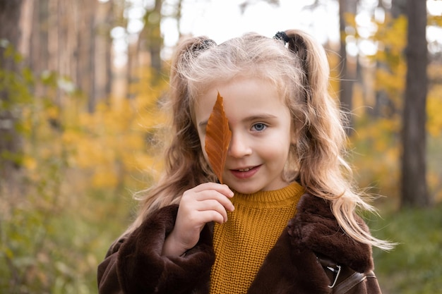 Happy little child girl smiling, covering her face with autumn leave in the fall forest