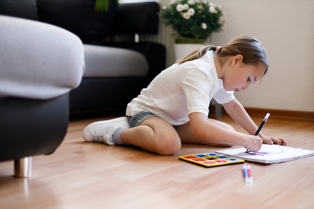 Happy little child is sitting comfortably on the wooden floor, drawing on paper with colored pencils