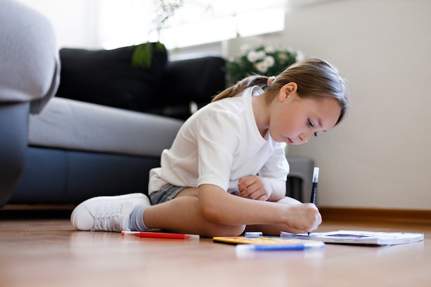 Happy little child is sitting comfortably on the wooden floor, drawing on paper with colored pencils