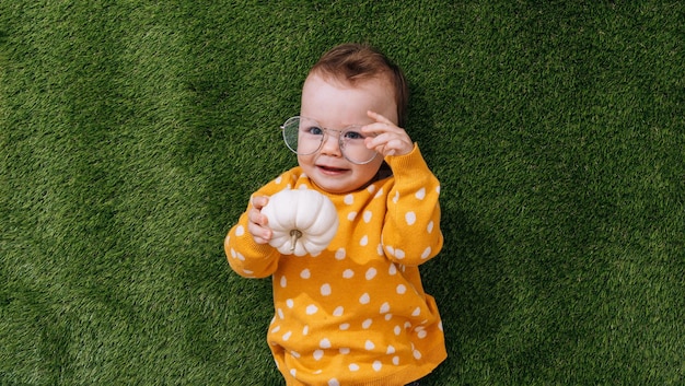 A happy little child lies on the green grass in a yellow sweater and glasses holds a pumpkin in his hands