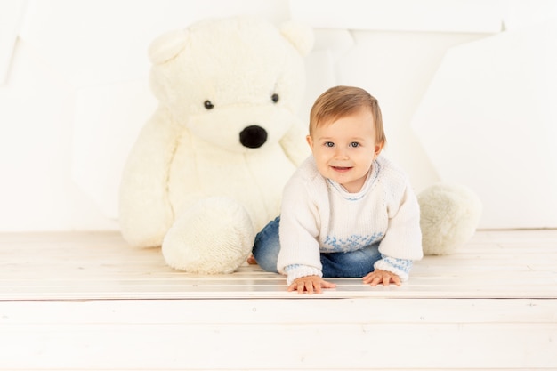 A happy little child six months old in a knitted warm jacket and blue jeans crawls at home near a large Teddy bear