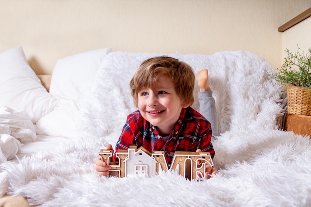 Happy little curly kid boy with smiley face lying on the bed at home Stay at home quarantine coronavirus COVID19 pandemic prevention Wooden house inscription home