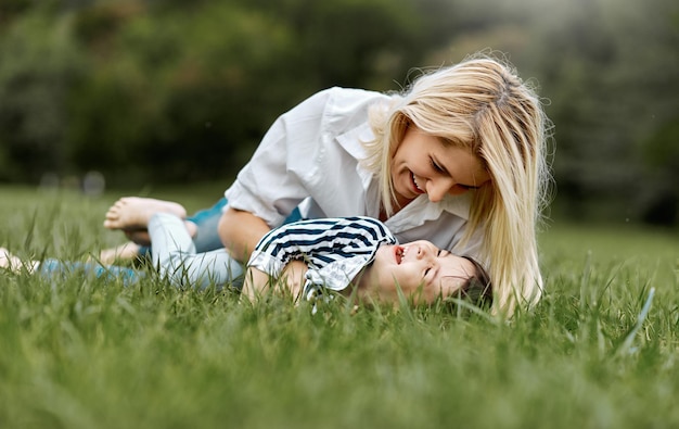 Happy little daughter lying on the green grass and playing with her smiling mother in the park Loving woman and her little kid girl enjoying the holiday Mom and child has fun outdoors Mothers day