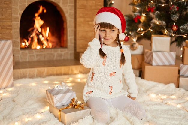 Happy little girl in beautiful white sweater and red santa hat in front of New Year Christmas tree