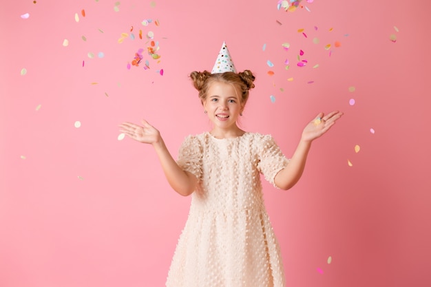 Happy little girl in a birthday cap blows off her palms multicolored confetti on a pink background in the studio