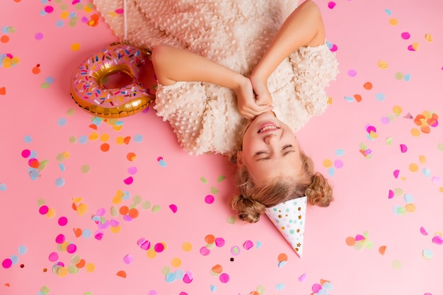 Happy little girl in a birthday cap lies among the multicolored confetti on a pink background