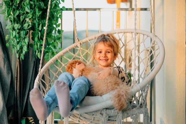 Happy little girl child hugging with a smile her pet poodle dog at home on the balcony in spring summer in a cottonfringed hammock chair at sunset