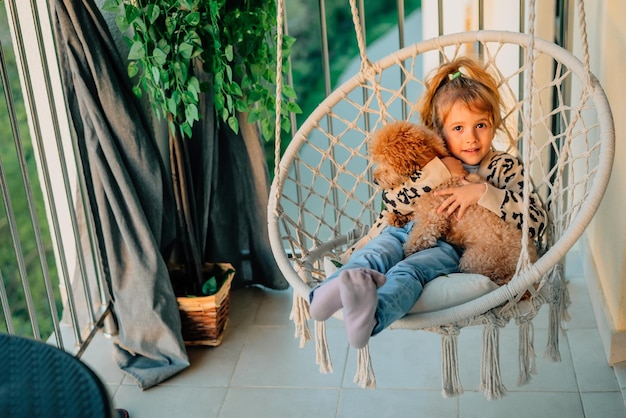 Happy little girl child hugging with a smile her pet poodle dog at home on the balcony in spring summer in a cottonfringed hammock chair at sunset