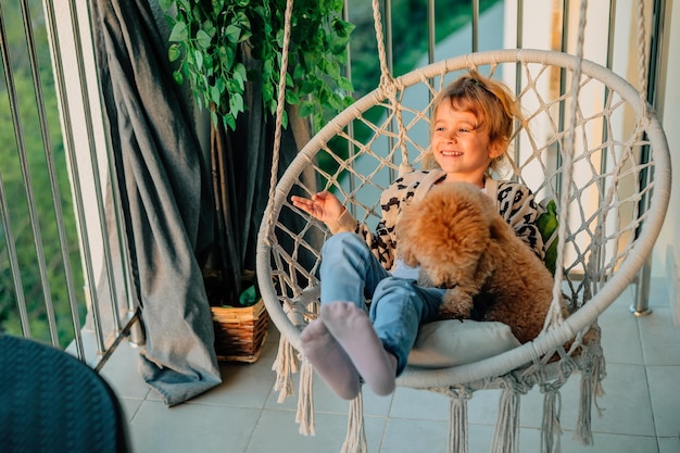 Happy little girl child hugging with a smile her pet poodle dog at home on the balcony in spring