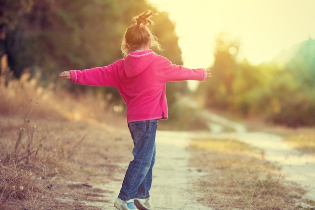 Happy little girl dancing on dirt rural road in summer back to camera