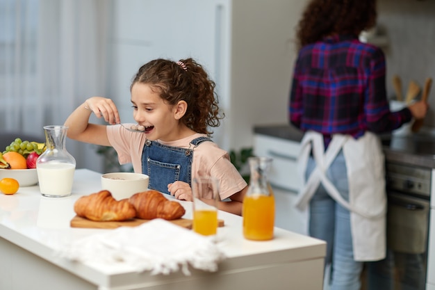 The happy little girl eating cereals healthy breakfast together with mother in the kitchen.