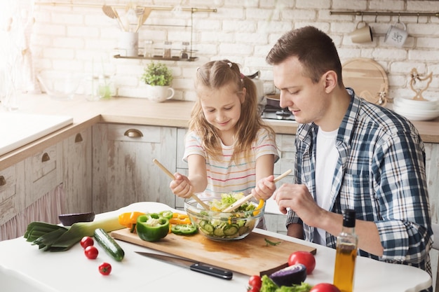 Happy little girl and her dad cooking at home kitchen. Father and daughter making vegetable salad and having fun together. Fathers day, family and healthy food concept