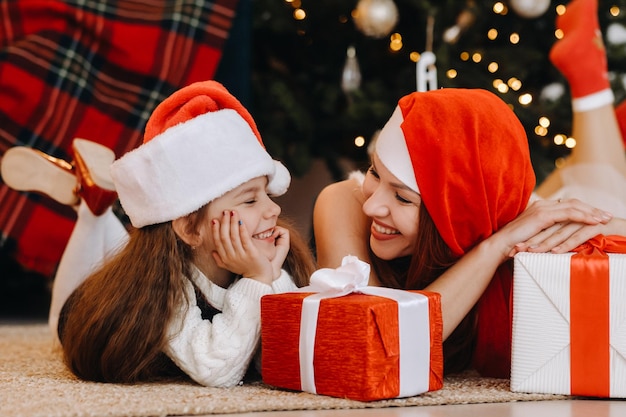 A happy little girl and her mom in Santa Claus hats are smiling with gifts in their hands.