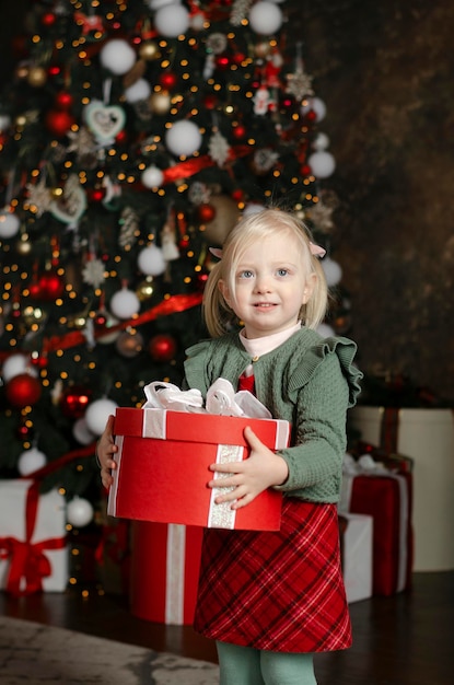 Happy little girl holds large big gift box and looks into camera on xmas tree background Christmas Eve Vertical frame