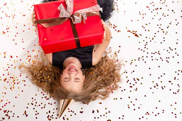 A happy little girl lies on a white background with a gift box and a cap for her birthday.