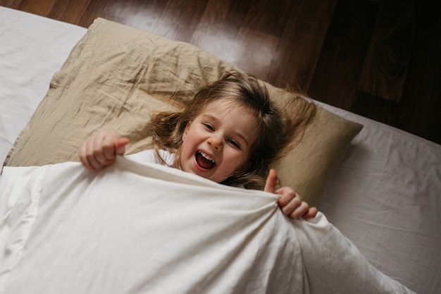 Happy little girl lying in bed, hiding under blanket