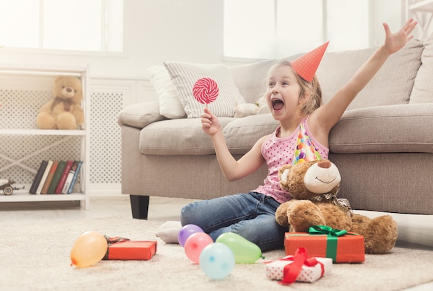 Happy little girl in party hat celebrating birthday. Smiling kid sitting on floor among gifts and balloons, holding lollipop. Holiday concept, copy space