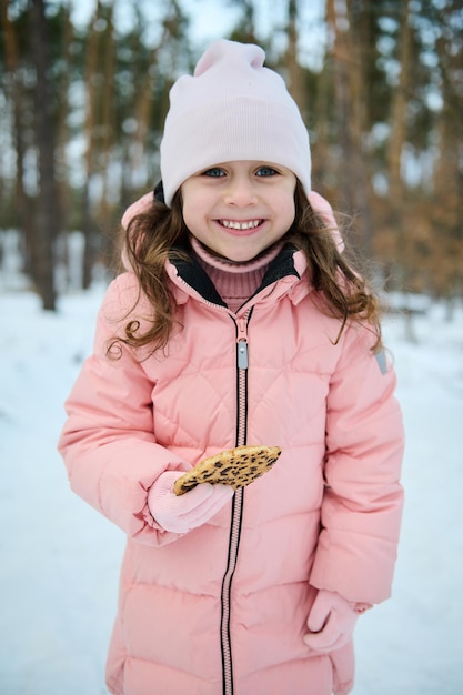 Photo happy little girl in pink pastel warm winter clothes holds cookies in her hand and smiles with a toothy smile looks at the camera while playing in a snowy forest enjoy the wonderful winter outdoors