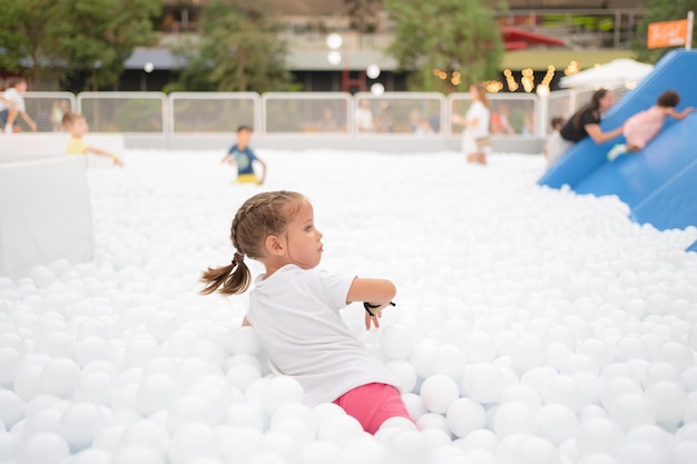 Happy little girl playing white plastic balls pool in amusement park playground for kids