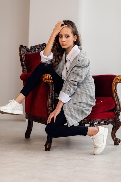 Happy little girl portrait little girl in a gray jacket in a crooked interior with a burgundy armchair white sneakers childrens fashion happy kids girl model