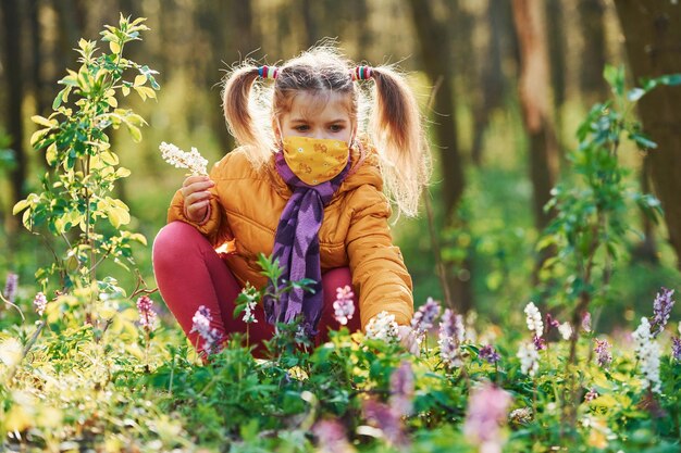 Happy little girl in protective mask sitting in spring forest at daytime