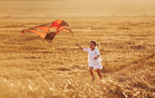 Happy little girl running with red kite outdoors on the field at summertime