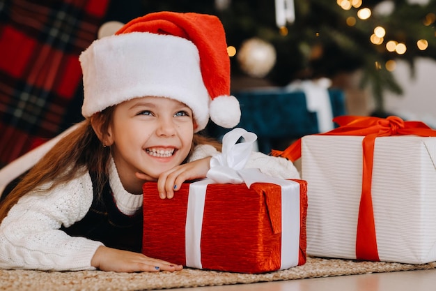 A happy little girl in a Santa Claus hat smiles with gifts in her hands.