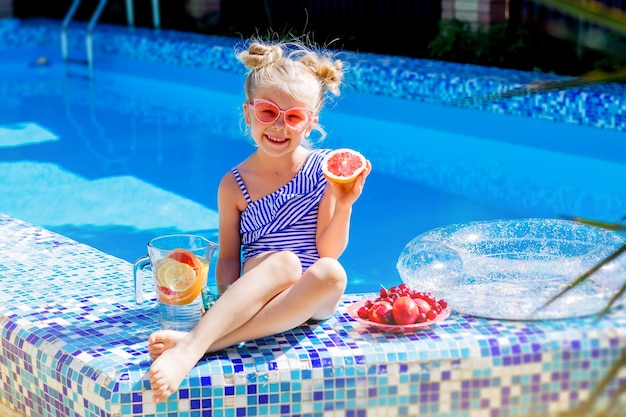 Happy little girl sitting by the pool in the summer drinking lemonade