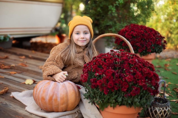 Happy little girl sitting on porch of house with chrysanthemums potted and pumpkins on Halloween