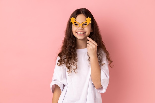 Happy little girl smiling and holding paper glasses on stick using party props on holiday having fun