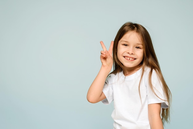 Happy little girl smiling showing V peace sign hand gesture on blue background Peace to the world stop the war